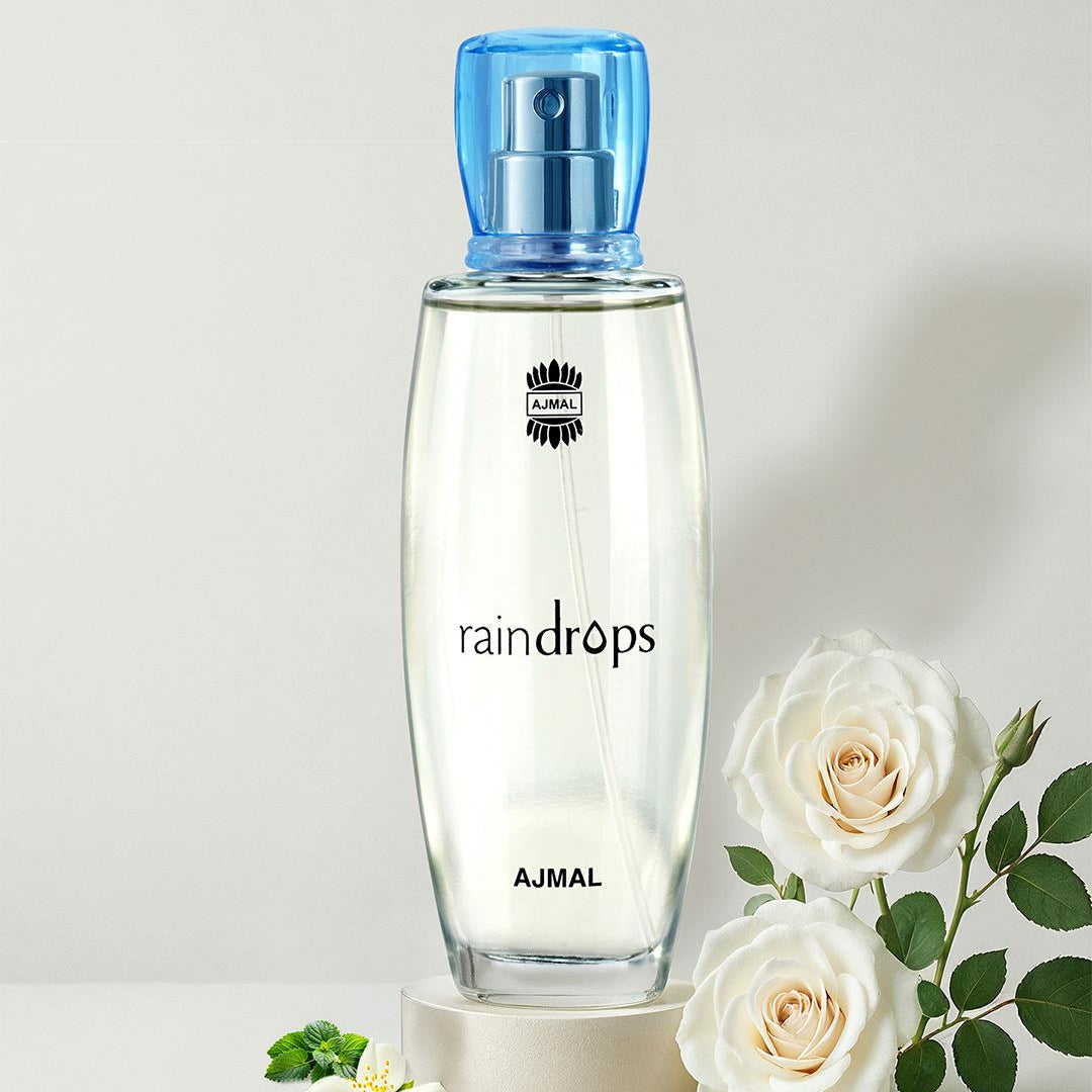 A clear glass bottle of raindrops AJMAL perfume with a translucent blue spray cap, displayed on a small beige pedestal with white roses and green leaves on a light gray background.
