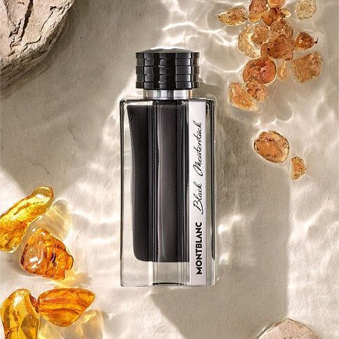 Montblanc Blanc Chantilly Black perfume bottle with a black cap, set on a textured beige surface with water ripples, amber stones, and natural rocks.